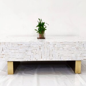 May include: White coffee table with a geometric pattern made of mother of pearl inlay. The table has a rectangular top and four gold metal legs.