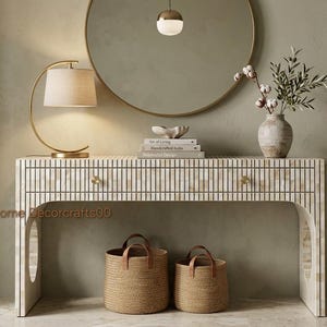 May include: A rectangular console table featuring a vertical striped pattern of off-white and beige tiles, two drawers with small gold knobs, and arched side cutouts. Two woven baskets with brown leather handles sit on the floor beneath the table.