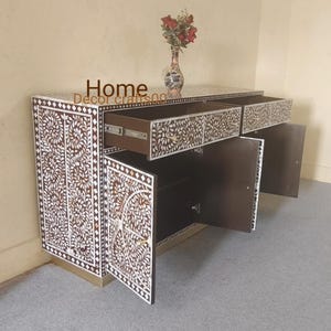 Puede incluir: Un armario de madera marrón con tres cajones y dos puertas. El armario está decorado con intrincados patrones florales blancos hechos de nácar. El armario está sobre una superficie marrón claro.