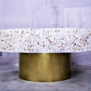 Pode incluir: Uma mesa redonda branca com uma base dourada. A superfície da mesa é decorada com um padrão de folhas marrons e brancas.