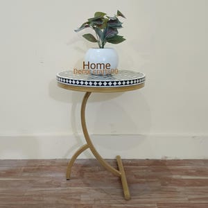 May include: A round side table with a white and black bone inlay top and a gold metal base. The table has a curved leg design and is decorated with a black and white geometric pattern. The table is topped with a white planter with a green plant.