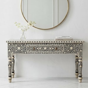 Può includere: Un tavolo consolle con motivo floreale in bianco e nero con un cassetto singolo e quattro gambe. Il tavolo ha un pomello dorato ed è sormontato da libri e un vaso di fiori. Uno specchio con cornice dorata è appeso sullo sfondo.