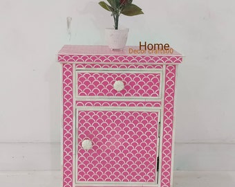 May include: A pink and white nightstand with a drawer and a door. The nightstand has a decorative pattern of white scales on a pink background. The nightstand is made of wood and has a white finish. The nightstand is 24 inches tall and 16 inches wide.