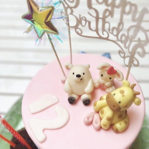 Könnte beinhalten: Eine zweistöckige rosa Torte, verziert mit einem weißen Eisbären, einer Giraffe und einem Hasen. Die Torte ist mit einem goldenen Glitzerstern und einem goldenen 'Happy Birthday'-Banner verziert. Die Torte ist von Schokoladen-Cupcakes mit Kerzen umgeben.