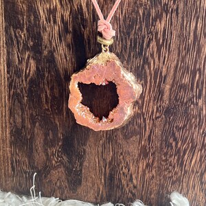 May include: A pendant necklace featuring a raw, rose-colored geode with a gold-colored trim. The geode is suspended from a light pink cord. The necklace is displayed against a dark wood background.