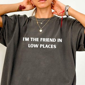 Model wearing a pepper Comfort Colors oversized t-shirt with white text reading “I’m the Friend in Low Places,” relaxed casual fit