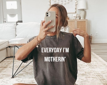 May include: Pepper-colored t-shirt with the text "EVERYDAY I'M MOTHERIN'" in white. The shirt is a relaxed fit, with a crew neck. The image also shows white socks with black stripes.