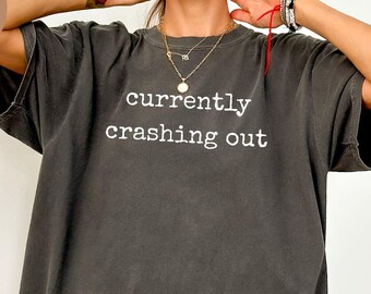 May include: A dark gray, oversized t-shirt with the words "currently crashing out" printed in white. The shirt is a pepper color and has a relaxed fit. The tee is a casual wear item.