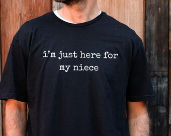 May include: Black t-shirt with the white text "i'm just here for my niece". The shirt is a crew neck style and is being worn by a person. The background is a wooden wall.