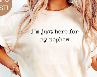 May include: Ivory-colored t-shirt with the text "i'm just here for my nephew" in black. The shirt is paired with blue jeans. The image also includes a gold bracelet and a gold necklace.