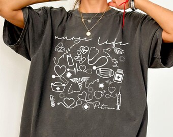 May include: Pepper-colored t-shirt with a white graphic design featuring medical symbols and the words "nurse life" and "patients". The design includes stethoscopes, hearts, and medical crosses. The shirt has short sleeves and a relaxed fit.