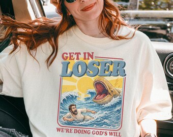 May include: Ivory-colored graphic t-shirt with the text "GET IN LOSER" above a retro illustration of a person in the ocean being chased by a shark. The shirt also has the text "WE'RE DOING GOD'S WILL".