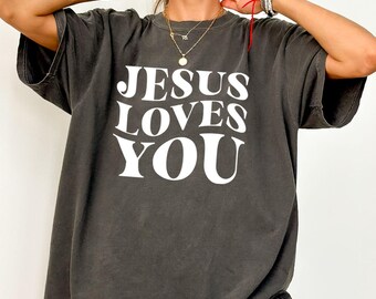 May include: A dark gray t-shirt with the text "JESUS LOVES YOU" in large white letters. The shirt has short sleeves and a relaxed fit. The color is described as "pepper".