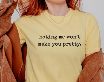 May include: A butter-colored t-shirt with the text "hating me won't make you pretty" in black. The shirt is paired with a brown jacket. The image is well-lit and focuses on the shirt's message.