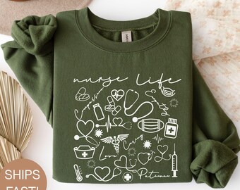 May include: Military green sweatshirt with the words "nurse life" and various medical-related icons in white. The design includes stethoscopes, hearts, and medical symbols. The sweatshirt is folded, with the sleeves rolled up.