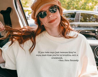 May include: Ivory-colored t-shirt with the quote "If you hate cops just because they're cops, next time you're in trouble, call a crackhead. -Sen. John Kennedy" printed in black. The shirt is worn by a person in a car.