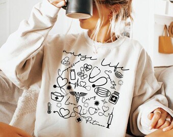 May include: Sand-colored sweatshirt with a black graphic design featuring medical symbols and the words "Nurse Life." The sweatshirt is paired with denim shorts. A black mug is held up to the face.