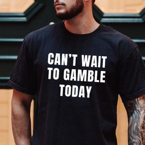 May include: A black t-shirt with white text that reads "Can't wait to gamble today". The shirt is being worn by a person with tattoos on their arm.