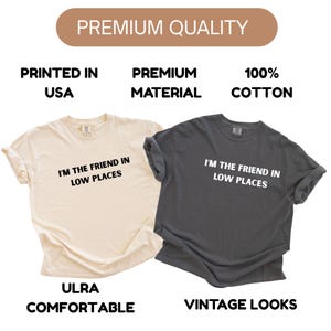 May include: Two t-shirts, one cream-colored and one charcoal gray, are displayed. Both shirts have the text "I'M THE FRIEND IN LOW PLACES" printed in white. The cream shirt has the words "ULRA COMFORTABLE" printed below the text, and the gray shirt has "VINTAGE LOOKS".
