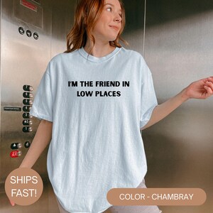 Model wearing a chambray Comfort Colors oversized t-shirt with white text reading “I’m the Friend in Low Places,” relaxed casual fit