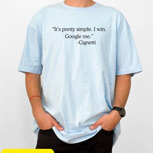 May include: Light blue t-shirt with the quote "It's pretty simple. I win. Google me." attributed to Cignetti in black text. The shirt is worn by a person with their hands in their pockets. The words "Sport Grey" are in a yellow rectangle.