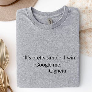 May include: Sport grey t-shirt with the text "It's pretty simple. I win. Google me." and the name "-Cignetti" in black. The shirt is folded and has a Gildan tag.