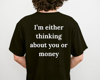 May include: A black short-sleeved t-shirt featuring white text printed on the back that reads, I'm either thinking about you or money. The shirt has a crew neckline and a standard fit, displaying the text clearly against the dark fabric background.