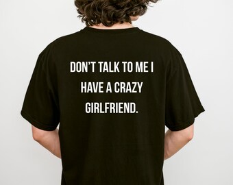 May include: A black short-sleeved t-shirt featuring white text printed on the back that reads, DON'T TALK TO ME I HAVE A CRAZY GIRLFRIEND. The shirt has a crew neckline and a standard fit, displaying the message clearly against the dark fabric.