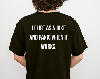 May include: A black short-sleeved t-shirt featuring white text on the back that reads: I FLIRT AS A JOKE AND PANIC WHEN IT WORKS. The shirt has a crew neckline and a standard fit, displaying the printed message clearly against the dark fabric background.