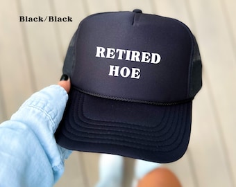 May include: Black trucker hat with the words "RETIRED HOE" in white block letters. The hat has a black brim and mesh back. The text "Black/Black" is in the top left corner.