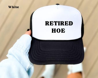 May include: A black and white trucker hat with the text "RETIRED HOE" in bold, black letters. The hat has a white front panel and a black brim and mesh back. The word "White" is printed in the upper left corner.
