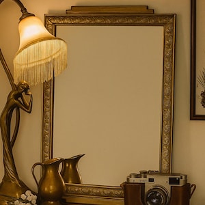 Peut inclure: Une scène de style vintage avec un miroir doré, une lampe en laiton avec un abat-jour à franges et un pichet en laiton. Un appareil photo dans un étui en cuir repose sur une surface en bois, créant une ambiance chaleureuse et nostalgique.