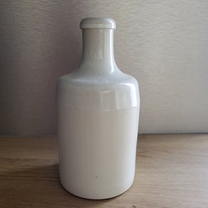 Peut inclure: Une bouteille en céramique blanche avec un corps arrondi et un col étroit. La bouteille est posée sur une surface en bois.