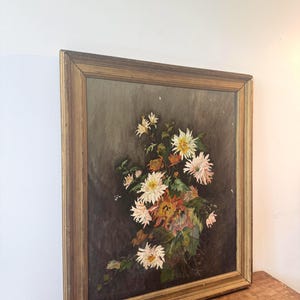Op de afbeelding: Een antiek bloemenschilderij in een houten lijst. Het kunstwerk toont een boeket witte, roze en oranje chrysanten tegen een donkere achtergrond. De lijst heeft een warme, goudbruine kleur, wat bijdraagt aan de vintage esthetiek.