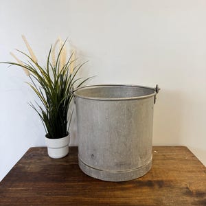 May include: A silver metal bucket with a handle on the side. The bucket is sitting on a wooden table with a potted plant in the background.
