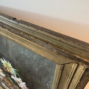 Op de afbeelding: Een antiek olieverfschilderij met een bloemstilleven met witte en gele bloemen tegen een donkere achtergrond. Het schilderij is ingelijst in een verweerde, goudkleurige houten lijst, met tekenen van ouderdom en slijtage.