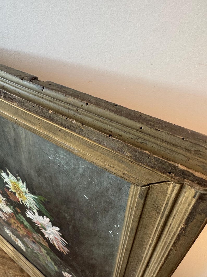 Op de afbeelding: Een antiek olieverfschilderij met een bloemstilleven met witte en gele bloemen tegen een donkere achtergrond. Het schilderij is ingelijst in een verweerde, goudkleurige houten lijst, met tekenen van ouderdom en slijtage.