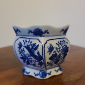 Peut inclure: Petit pot en céramique blanche avec un motif floral bleu. Le pot a un bord festonné et une base en piédestal. Le motif floral bleu est répété autour de l'extérieur du pot.