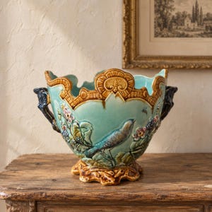 Może przedstawiać: Ceramiczna donica z jasnoniebiesko-zielonym szkliwem i wypukłymi detalami przedstawiającymi ptaka, kwiaty oraz liście. Krawędź i podstawa mają brązowe zdobienia, a naczynie posiada dwa ciemne uchwyty po bokach.