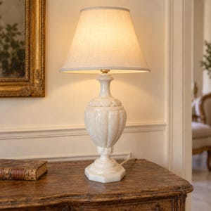 May include: A table lamp with a cream-colored, urn-shaped base made of polished stone featuring vertical fluted detailing and a square pedestal. The lamp is topped with a tapered, off-white fabric lampshade that emits a warm, soft glow.
