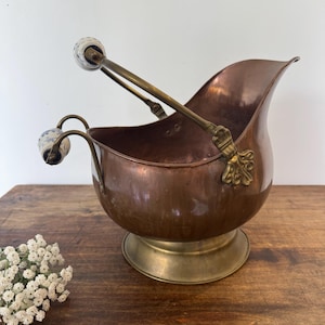 Peut inclure: Seau à charbon en cuivre avec une base et une poignée en laiton. La poignée a des extrémités décoratives en céramique avec des motifs bleus et blancs. Le seau a une forme incurvée et est conçu pour contenir du charbon. Le seau est posé sur une surface en bois.