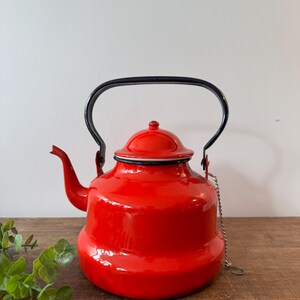 Peut inclure: Une bouilloire en émail rouge vif avec une anse et un bec noirs. La bouilloire a un couvercle rouge assorti avec un bord noir et une petite chaîne attachée. La bouilloire est posée sur une surface en bois.