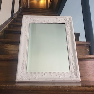 May include: A rectangular white framed mirror with ornate floral and beaded detailing. The mirror is resting on a wooden surface. The frame has a distressed, vintage appearance, adding to its charm. The mirror reflects the surrounding environment.