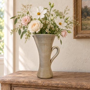 Puede incluir: Un jarrón de cerámica alto de color beige claro con borde acampanado y una sola asa lateral, lleno de un ramo de cosmos blancos, rosas de color rosa pálido y pequeñas flores verdes. El jarrón tiene un acabado liso y moteado con base cónica.