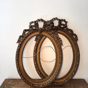 Op de afbeelding: Twee ovaalvormige fotolijsten met een metallic gouden afwerking. Elke lijst is voorzien van een sierlijk, verhoogd strikdetail aan de bovenzijde en een gestructureerd, decoratief randpatroon langs de binnen- en buitenranden van de ovale vorm.
