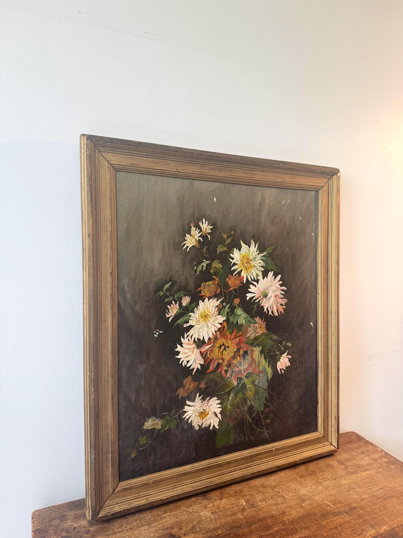 Op de afbeelding: Een antiek bloemenschilderij in een houten lijst. Het kunstwerk toont een boeket witte, roze en oranje chrysanten tegen een donkere achtergrond. De lijst heeft een warme, goudbruine kleur, wat bijdraagt aan de vintage esthetiek.