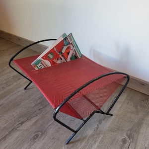 Peut inclure: Un porte-revues en fil rouge avec un cadre en métal noir. Le porte-revues est conçu avec une forme incurvée et a un magazine posé dessus.