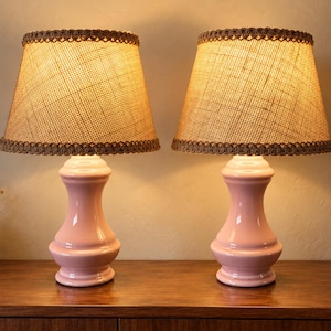 Puede incluir: Un par de lámparas de mesa con bases de cerámica rosa claro brillante y silueta curva en forma de reloj de arena. Cada lámpara tiene una pantalla cónica de arpillera beige texturizada con un borde trenzado marrón decorativo en los extremos.