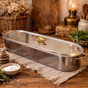 Könnte beinhalten: Ein Fischdämpfer aus Edelstahl mit einem goldfarbenen fischförmigen Griff. Der rechteckige Dämpfer hat goldene Griffe an jedem Ende. Der Dämpfer steht auf einem Holztisch mit einem Leinenläufer. Eine kleine Schale mit Salz und Kräutern befindet sich im Vordergrund.