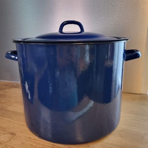 Pode incluir: Uma panela grande de esmalte azul com tampa a condizer. A panela tem duas pegas laterais e uma pega em laço na tampa. Está sobre uma superfície de madeira. A panela é azul escuro com um acabamento brilhante.
